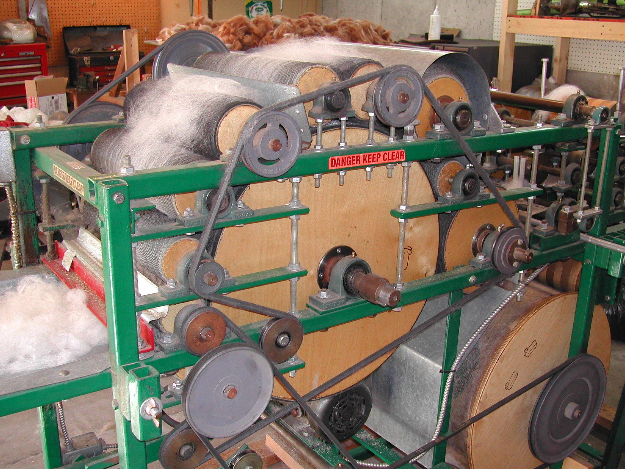 A crank-operated drum carder processing white alpaca fiber, showing the large main drum and small licker-in drum covered in fine wire teeth