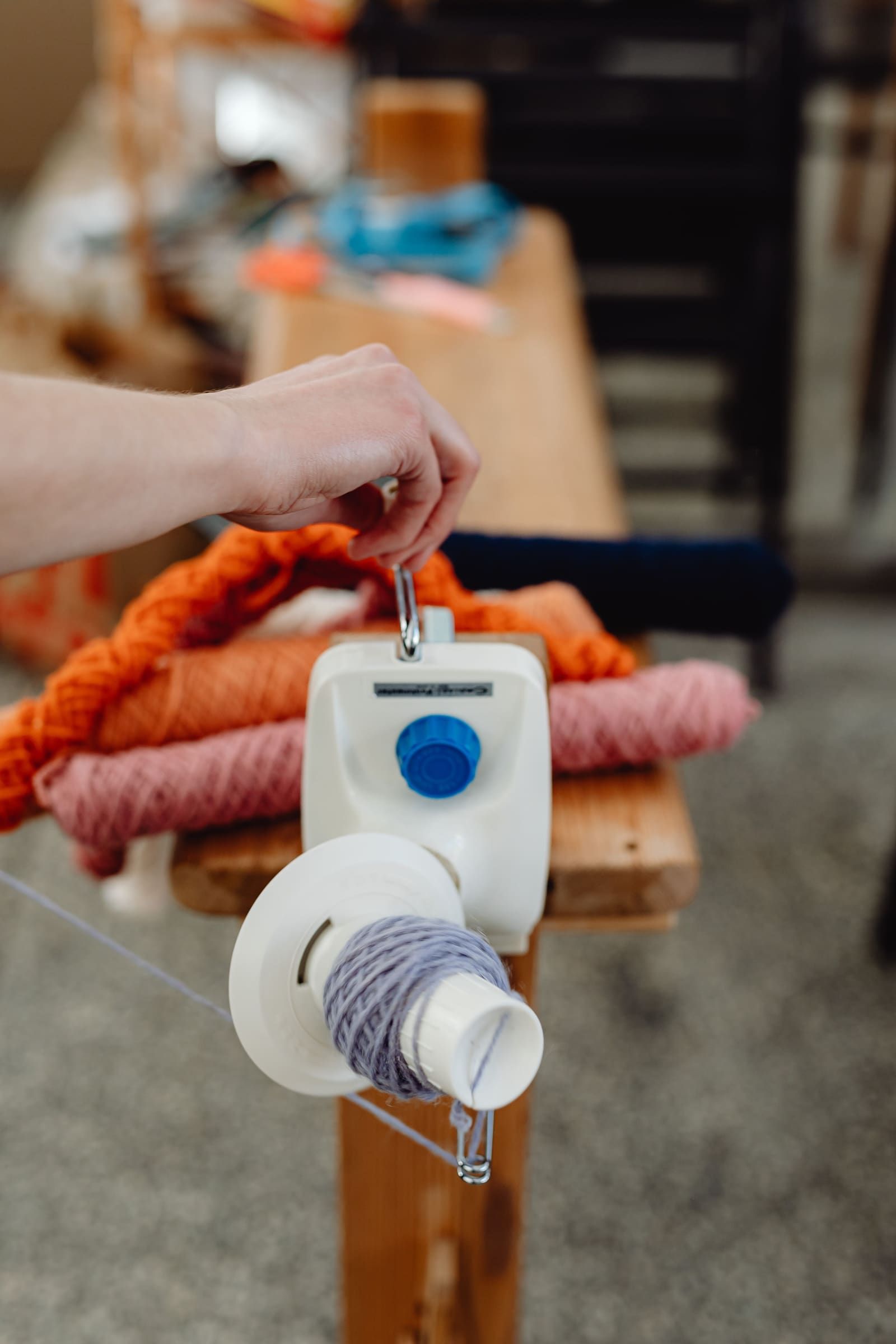 A hand lifts a ball winder's handle off its clamp mount, with loose hanks of orange and pink yarn coiled on the workbench behind it, ready to be wound