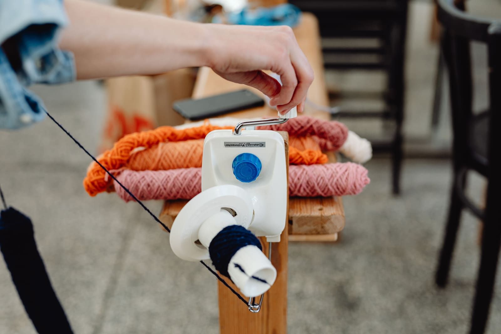 Close-up of a hand turning the crank of a plastic hand-crank ball winder, with a small center-pull cake building on the spindle and loose orange and pink yarn hanks visible on the table behind it