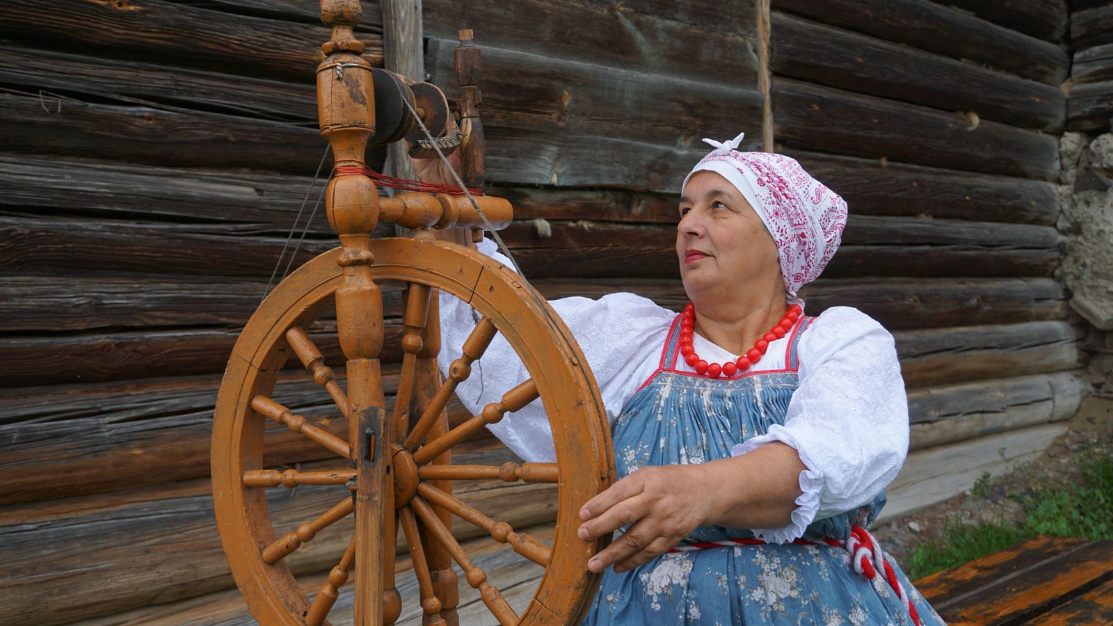 An elderly woman in traditional embroidered dress spinning at a Saxony-style spinning wheel outdoors, her hands drawing fiber through the orifice as the large drive wheel turns beside her