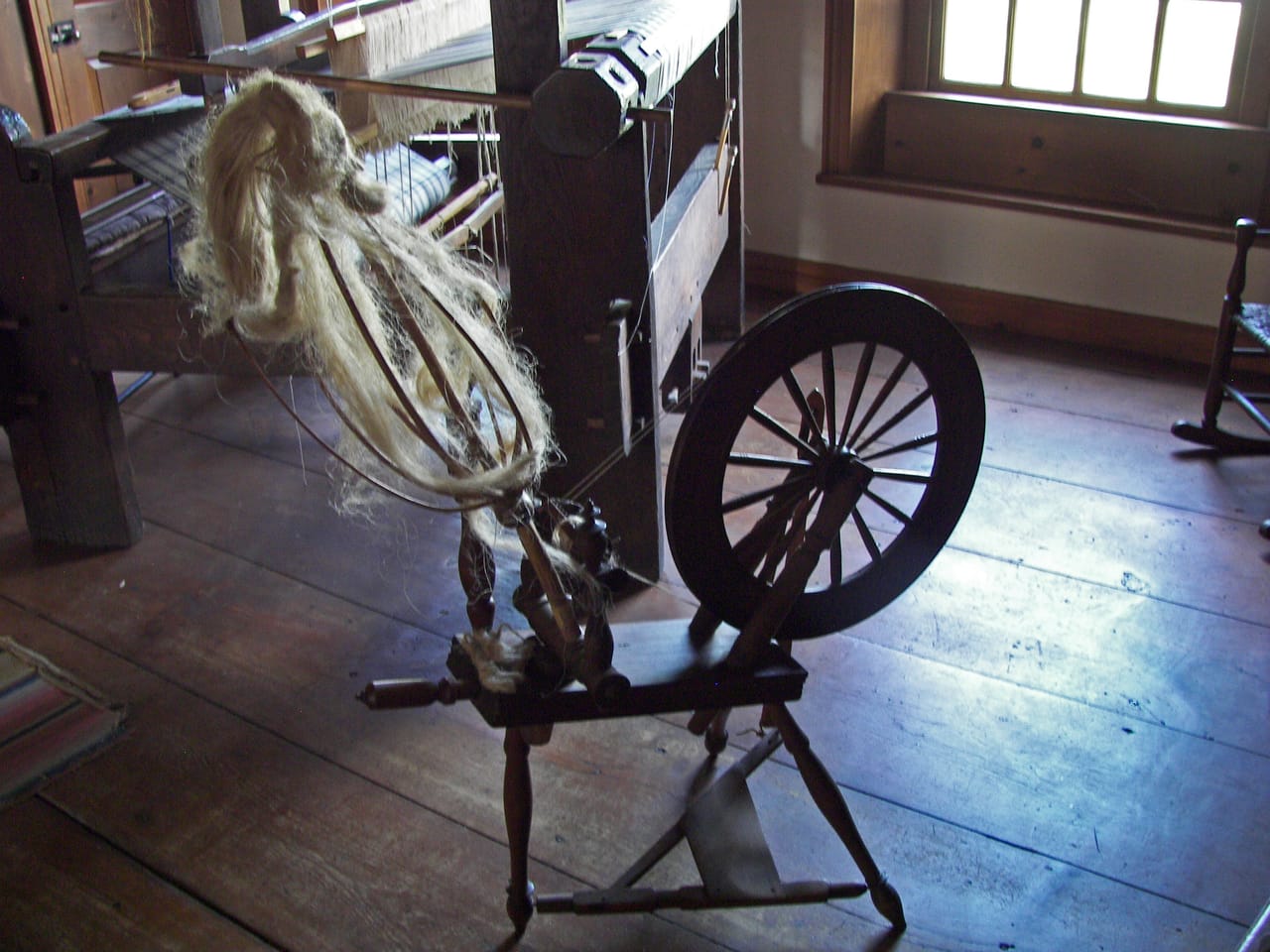 A Saxony spinning wheel displayed in a historic house museum, positioned in front of a floor loom, showing the large horizontal drive wheel and flyer-bobbin assembly arranged to one side on a wide, low frame