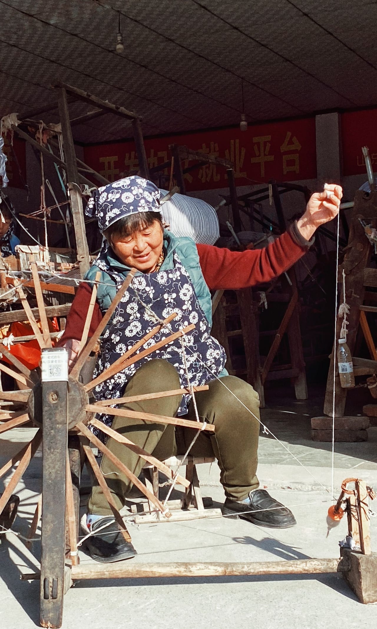 An elderly woman seated at a traditional spinning wheel indoors, her hands drawing out fiber as the wheel turns and the yarn builds on the bobbin, natural light falling across the frame and the spinner's face
