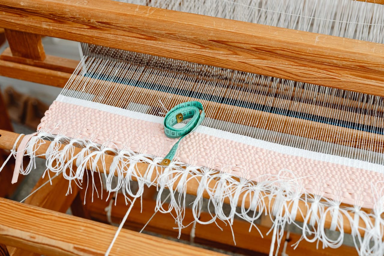 Several completed handwoven scarves in different colors folded on a wooden surface next to a small loom