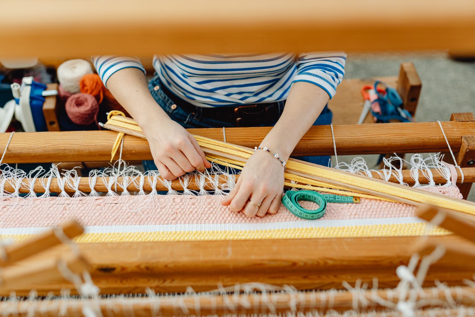 Hands weaving a striped table runner on a rigid heddle loom, warp threads in multiple colors