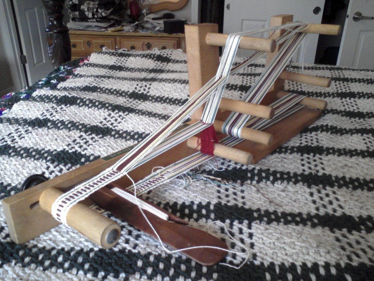 A table-model inkle loom with a white, pale green, marigold and brown band in progress, resting on a black and white woven cloth