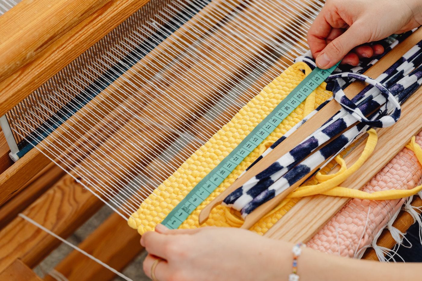 A weaver's hands holding a tape measure across a yellow woven strip on a wide loom with a pink strip rolled beside it
