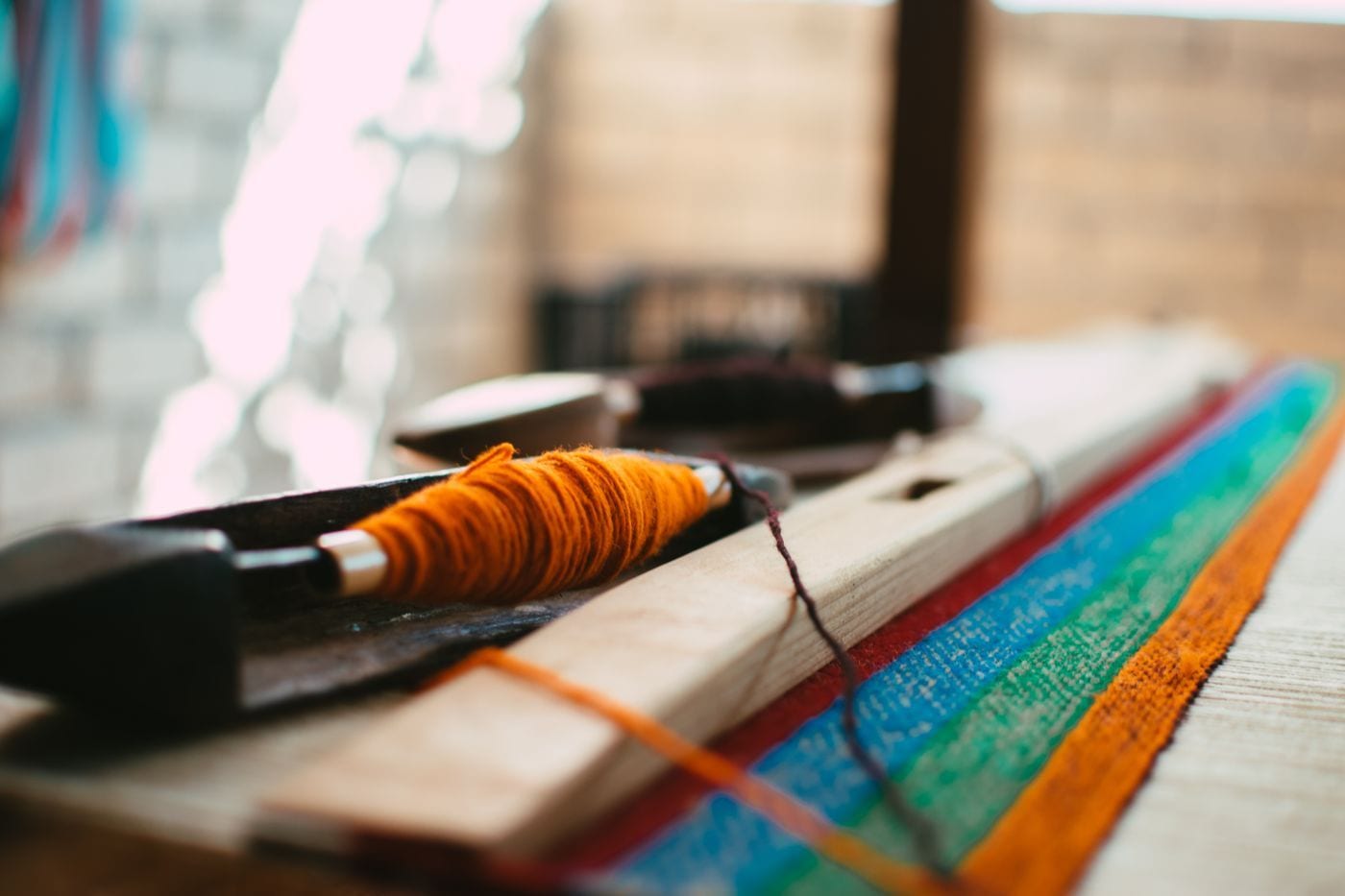 A wooden shuttle wound with orange wool lying beside a striped woven strip in red, blue, green and orange