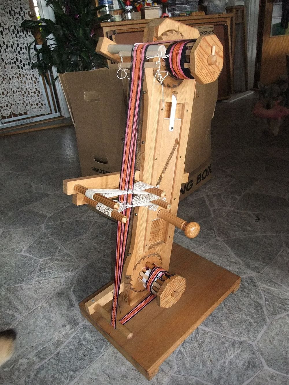 An upright wooden table-model loom with orange, pink and black warp threads running from a roller at the top past dowel pegs to a roller at the bottom, standing on a stone-tiled floor
