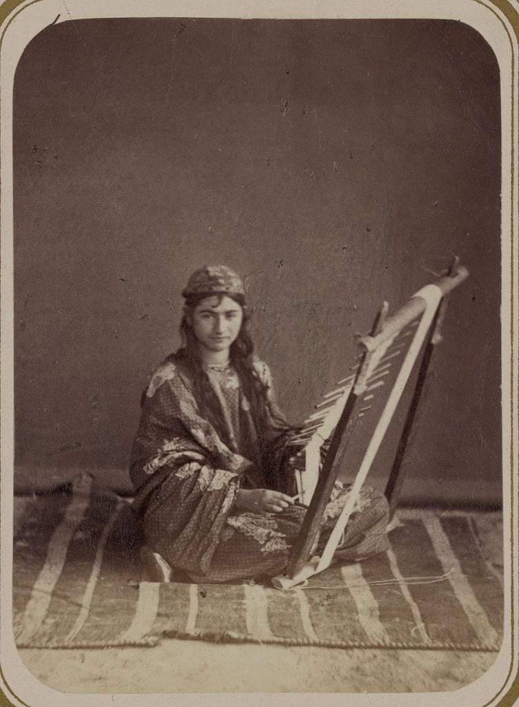 Sepia photograph of a woman in patterned dress and cap seated on a striped rug, working a small loom set at an angle in front of her