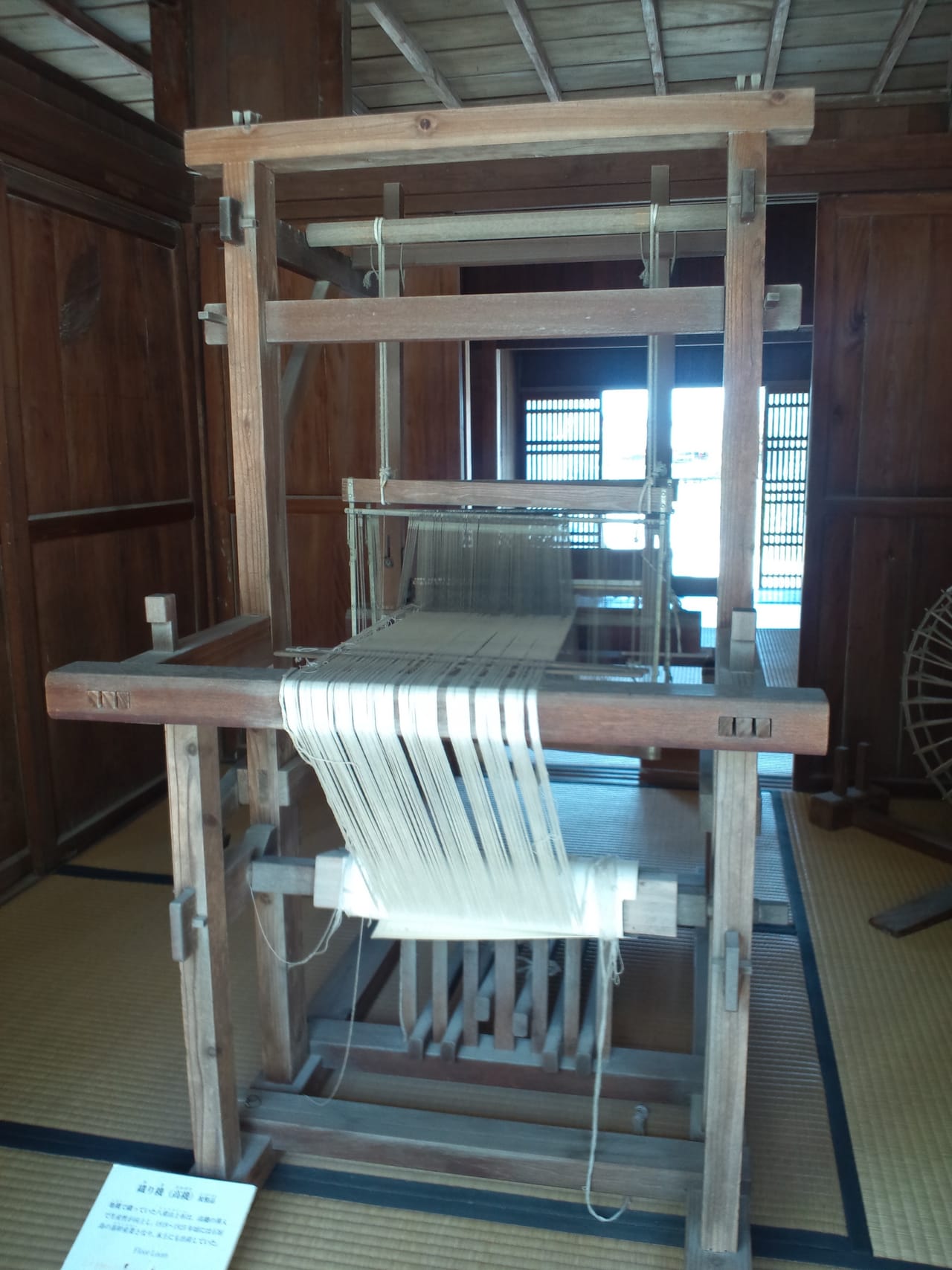 A traditional Japanese floor loom (takahata) displayed in a recreated house at an outdoor museum in Aichi Prefecture, showing the complete structure from warp beam at back to cloth beam at front with heddle frames and beater visible