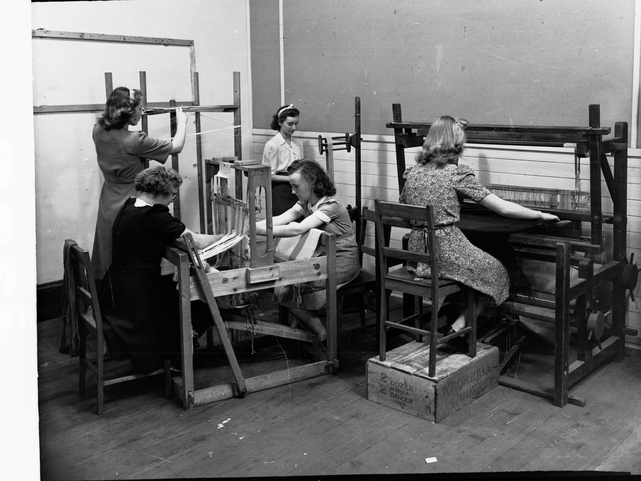 Young women weaving on looms in a school workshop setting, each at a separate loom, cloth building on the breast beams