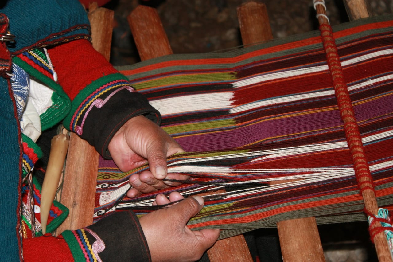 Overhead view of hands guiding a shuttle through a colorful warp on a wooden rigid heddle loom, showing the plain weave being formed row by row