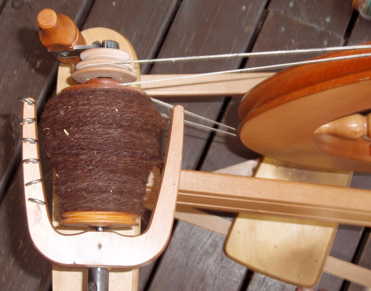 Close-up of a double-drive spinning wheel showing the drive band routed around both the flyer whorl and bobbin whorl with the flyer hooks visible